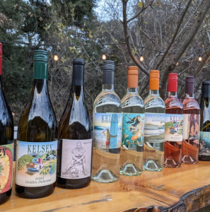 A row of Kelsey wine bottles with illustrated labels lined up on a live-edge wooden bar