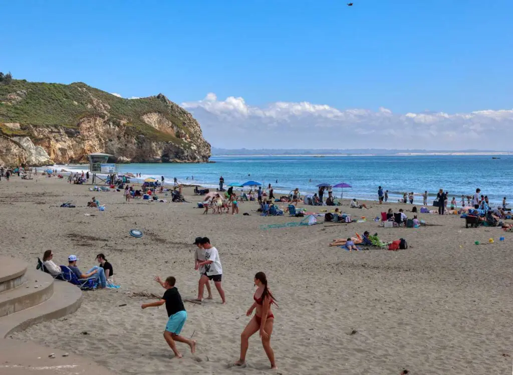A busy summer afternoon on Avila Beach, with umbrellas and towels along the shore below the headland cliff