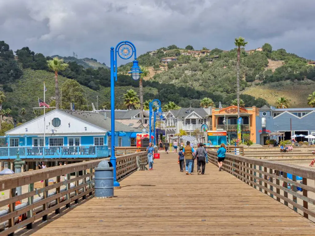 Looking along the wooden deck of the Avila Beach pier toward town, with blue lamp posts and people walking