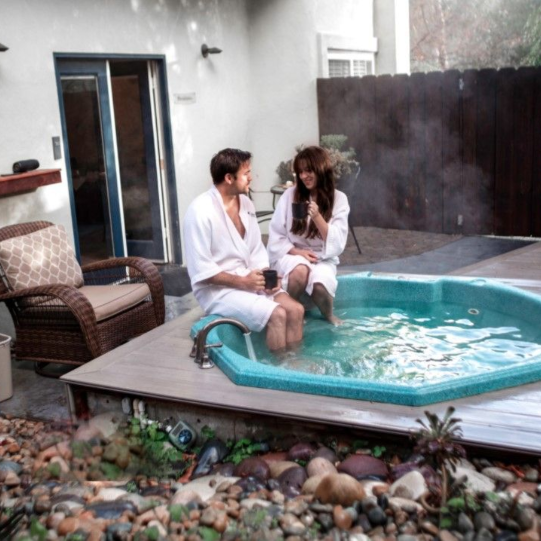 A couple in white robes sharing coffee on the edge of a private mineral spring hot tub outside a suite at Sycamore Mineral Springs