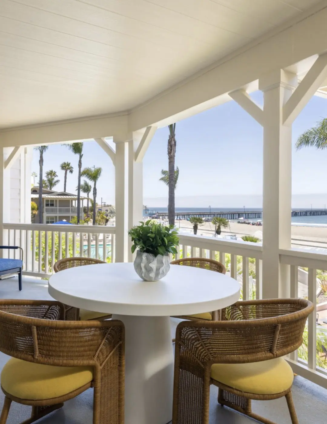 A suite balcony at Avila Beach House with wicker chairs and a round white table, looking past palm trees and the pool to the beach and the pier