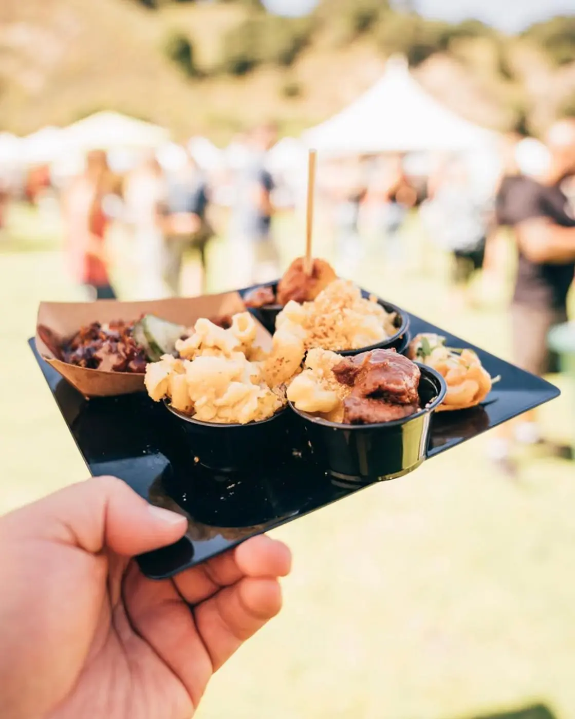 A hand holding a black tray of mac and cheese samples in small cups at the Avila Beach Mac and Cheese Festival, with festival tents and a grassy hillside behind