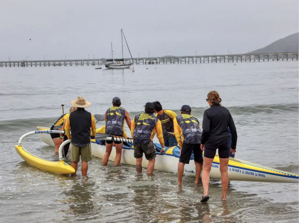 Paddlers walking a yellow and white outrigger canoe into the shallows at Avila Beach, with the pier behind