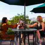 Three friends toasting with glasses of wine under green umbrellas on the garden patio at Avila Village Inn, with the hills behind