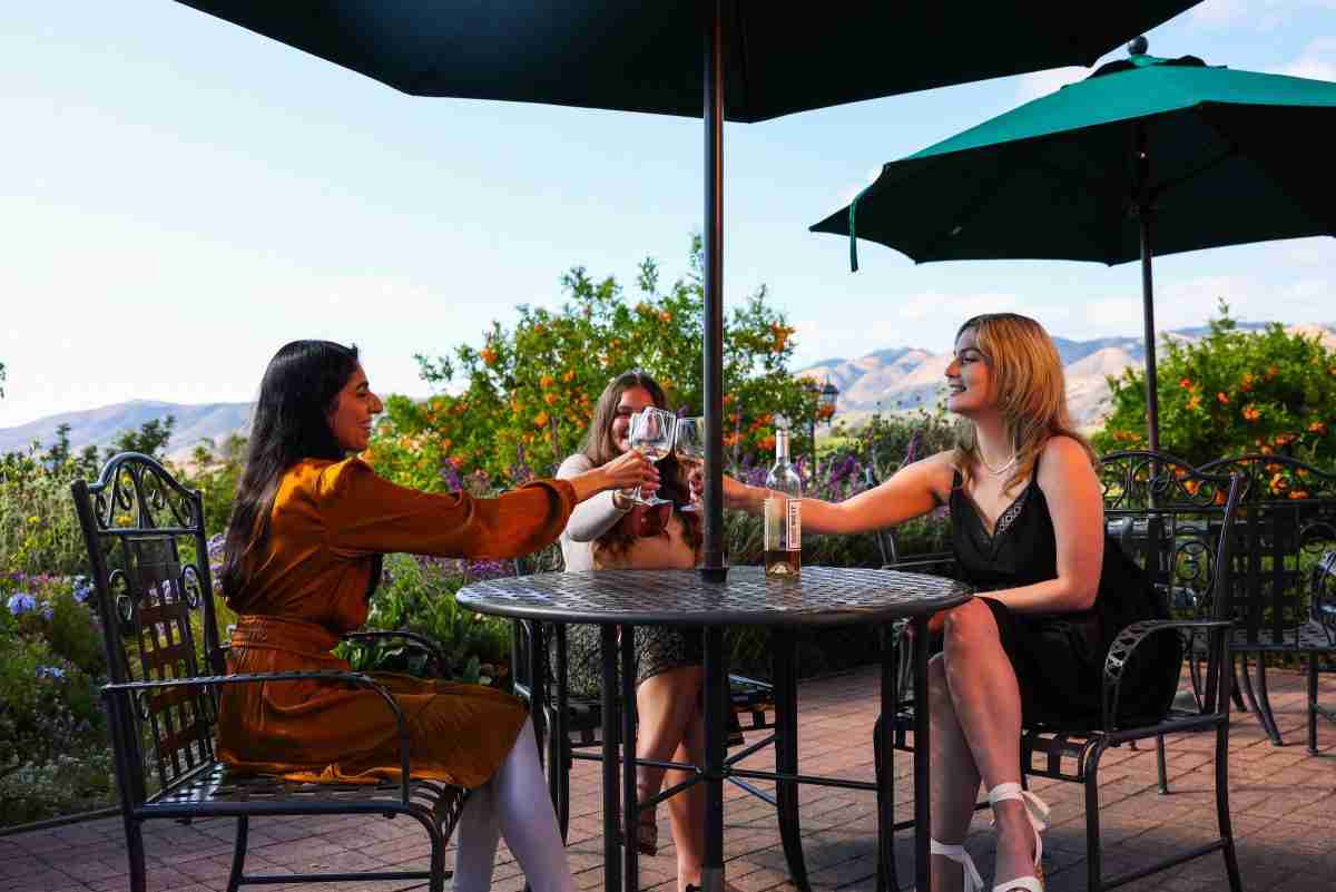 Three friends toasting with glasses of wine under green umbrellas on the garden patio at Avila Village Inn, with the hills behind